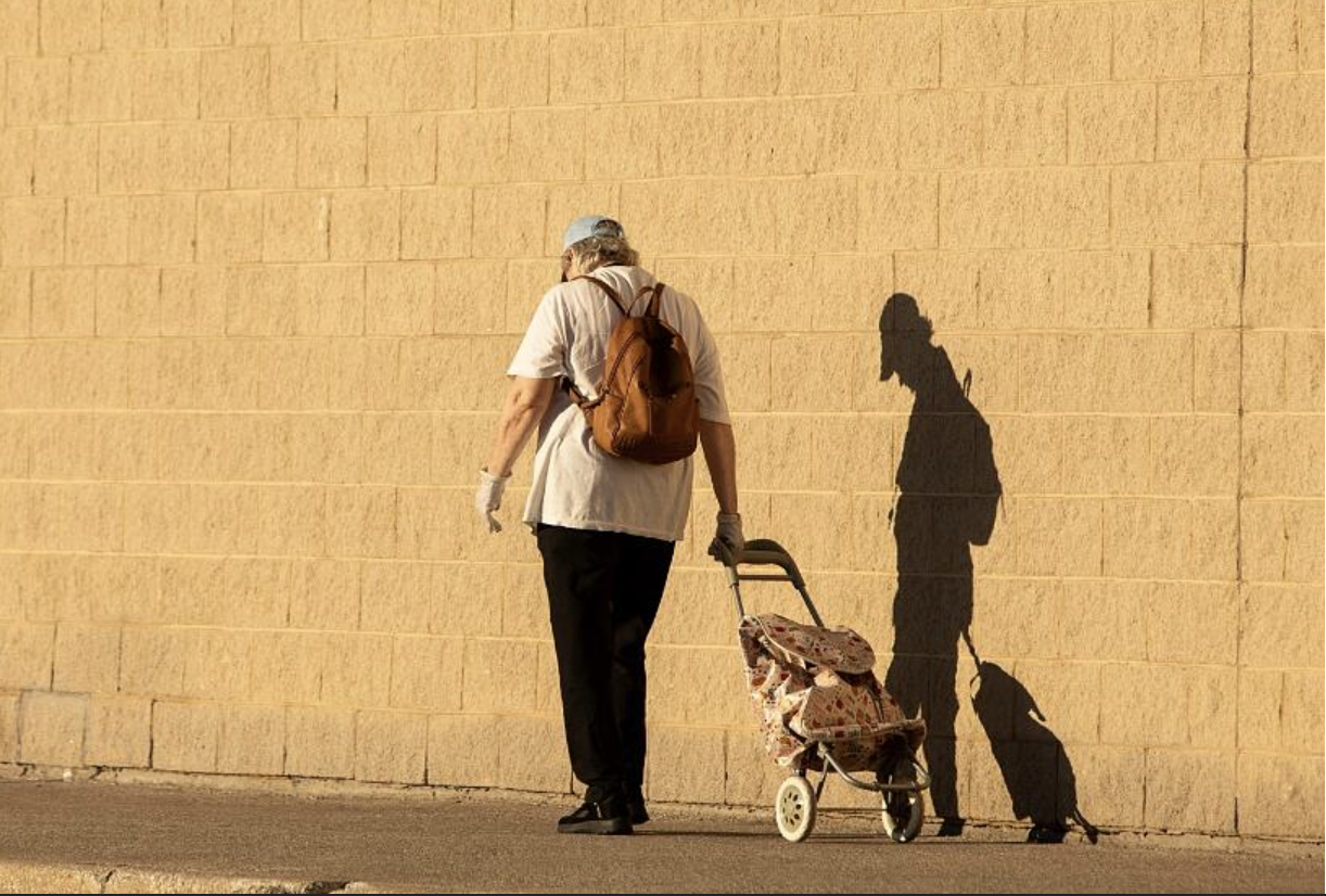 Person pulling a shopping trolley beside a sunlit yellow wall.