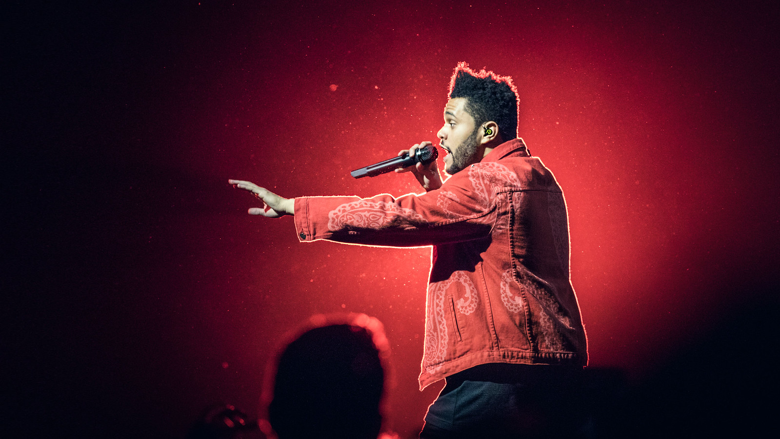 The Weeknd sings into a microphone onstage, wearing a red jacket under red concert lights.